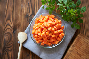 Cubes of carrots in a clear bowl on a gray cloth with a wooden spoon, top view. 