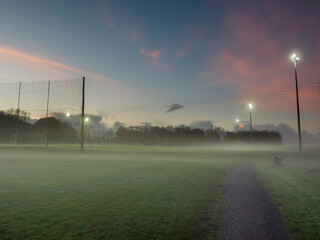 Fototapeta Irish National sport ground wit tall goal posts for camogie, hurling, rugby, Gaelic football at dusk and low fog over the ground. Calm and peaceful mood. Sport activity concept. Sunset cloudy sky. obraz