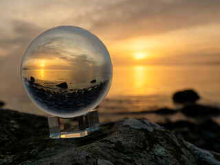 Glass ball on a rough stone by the ocean, sunset sky in the background. Warm soft light. Selective focus. Calm peaceful mood. Abstract nature scene.