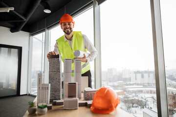 Qualified architect or engineer in reflective vest and hardhat inspecting skyscraper model in architectural modern office with panoramic city view. Bearded attractive man controlling working process.