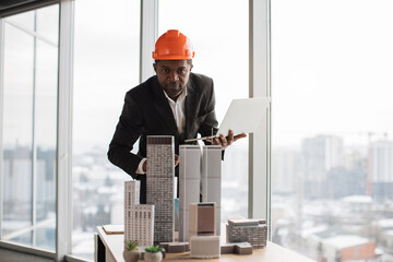 Focused african inspector using laptop inspecting skyscraper model in architectural modern office with panoramic city view on architectural bureau. Handsome man wearing suit and hardhat.