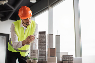 Bearded engineer in protective helmet working on building complex prototype project of residential or business district, measuring city model using compass for drawing at office with panoramic view