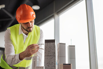 Bearded engineer in protective helmet working on building complex prototype project of residential or business district, measuring city model using compass for drawing at office with panoramic view