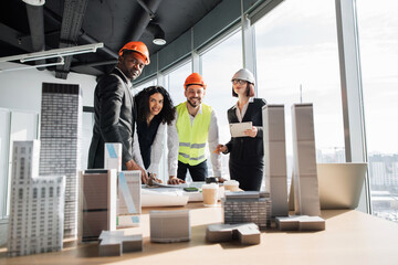 Fototapeta na wymiar Multiracial coworkers builders and architects standing near table with blueprints, gadgets and design of buildings residential project maquette and looking at camera.