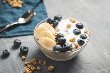 Granola cereal oatmeal with white yogurt, blueberries and banana fruits in a bowl on a blue napkin, grey background