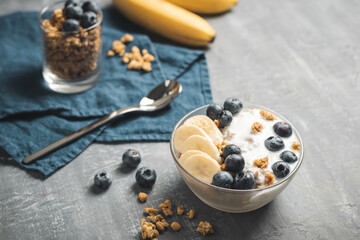 Granola cereal oatmeal with white yogurt, blueberries and banana fruits in a bowl on a blue napkin, grey background