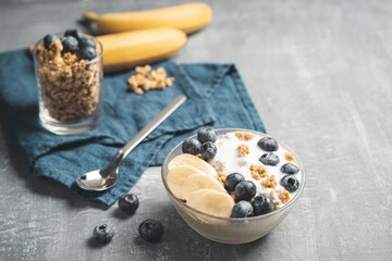 Granola cereal oatmeal with white yogurt, blueberries and banana fruits in a bowl on a blue napkin, grey background