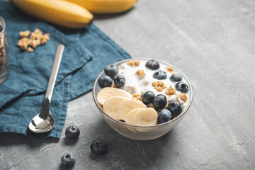 Granola cereal oatmeal with white yogurt, blueberries and banana fruits in a bowl on a blue napkin, grey background