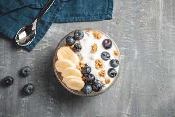 Granola cereal oatmeal with white yogurt, blueberries and banana fruits in a bowl on a blue napkin, grey background, top view