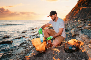 Earth Day. A male volunteer puts a plastic bottle in a bag full of garbage. In the background, the sea and the sunset. Copy space. The concept of environmental conservation