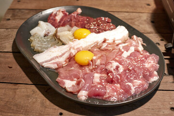 Set of meats for sukiyaki-shabu-yakiniku grill in a black plate placed on a wooden table. The raw meat set includes beef, pork, bacon, liver, fish, squid, shrimp and 2 eggs.