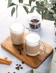 Two cappuccino or latte with milk foam in glasses on a wooden plate on a light background with coffee beans, cinnamon and eucalyptus branch.