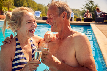 Retired Senior Couple Relaxing In Swimming Pool On Summer Vacation Celebrating Drinking Champagne