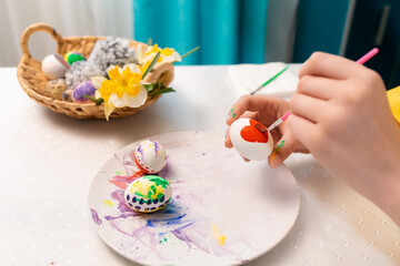 Close up of female hands decorating eggs for Easter. On the table there is a wicker basket with a toy rabbit and a daffodils. Concept of traditional celebrations and creativity
