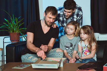 Staged photo. Homosexual couple and their children, two cute girls, at home.  An everyday evening after a day's work. Let's see what's in the big box! 