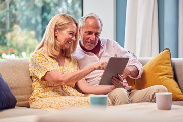 Senior Retired Couple Sitting On Sofa At Home Shopping Or Booking Holiday On Digital Tablet