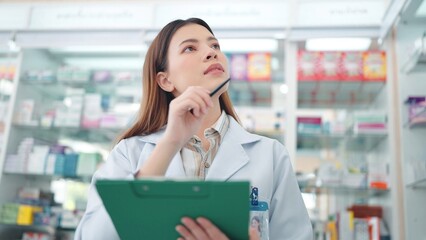 Professional Asian woman pharmacist rechecks products in pharmacy and using noteboard to write...