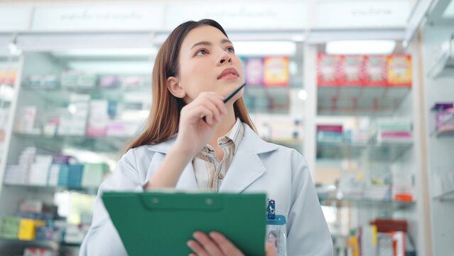 Professional Asian woman pharmacist rechecks products in pharmacy and using noteboard to write information