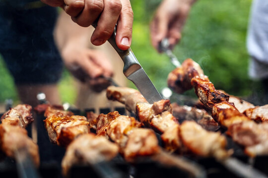 Man hands trying barbecue delicious grilled meat with knife preparing at the nature. Picnic with pork steaks on fired charcoals. Hot BBQ closeup