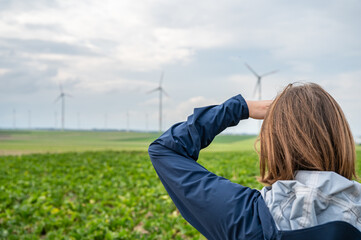 Woman with brown hair and blue jacket is looking at a wind park from the distance, wind park with...