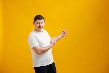 Handsome smiling man wearing white t-shirt standing pointing fingers aside on mock up copy space on yellow studio background