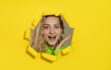 Surprised curly haired young lady have fun, poses in yellow paper hole for making photo, feels upbeat, expresses positive emotions. Wow face