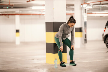 A tired sportswoman is leaning on the pillar and resting after workouts in garage.