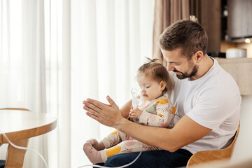 A father is holding his baby girl in his lap while she using nebulizer.