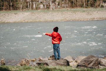 A little boy in a red jacket and jeans is playing on the shore and throwing stones into the river.