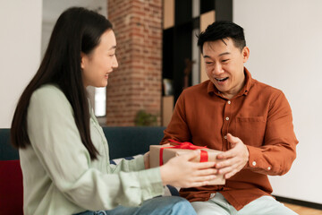 Loving young asian wife greeting her husband with marriage anniversary, giving him gift box and smiling