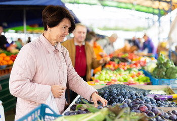 Positive mature woman in casual clothes picking fresh plums during shopping at grocery market