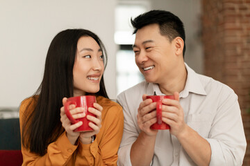 Happy loving japanese spouses sitting on sofa in living room and drinking hot beverage together, enjoying time at home