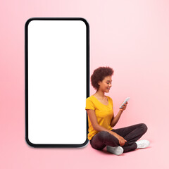 Smiling young black curly lady sit on floor with phone near big smartphone with empty screen