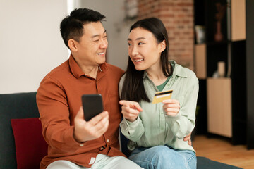 Banking application and e-commerce concept. Excited asian spouses using smartphone and credit card, sitting on sofa