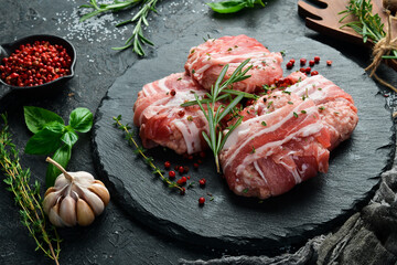 Raw cutlets wrapped in bacon served on a black plate with rosemary and thyme. On a stone...