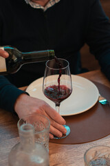 Camarero echando una copa de vino tinto a una copa de cristal sujetada por la mano de un hombre de raza europea encima de una mesa con platos