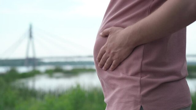Side view overweight Caucasian young man touching tummy standing on river bank on overcast day. Unrecognizable plus-size guy with beer tummy outdoors on spring summer day
