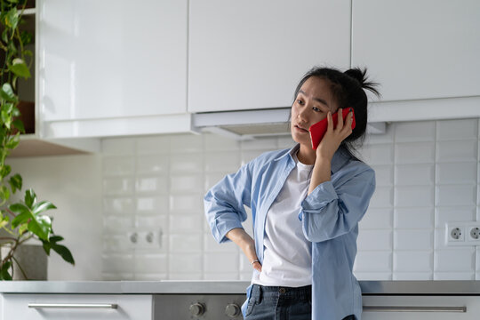 Young disappointed Asian woman standing in kitchen at home talking on smartphone hearing bad news. Stressed female holding mobile phone feeling upset during unpleasant phone conversation