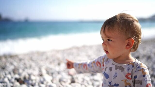 Little girl sits on a pebble beach, points to the surf and babbles
