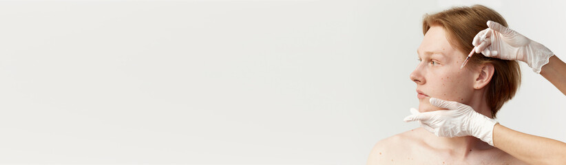 Young redhead man having cosmetological injections on cheekbone. Male model posing against grey...