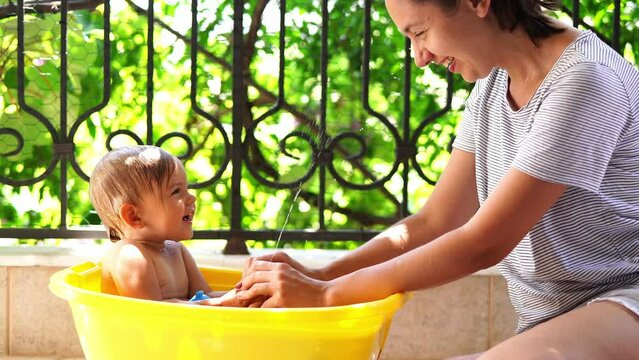 Little girl splashes water from a rubber duck on her mother, sitting in a basin on the balcony
