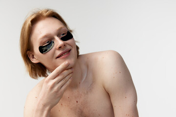 Young redhead man with eye patches posing shirtless against grey studio background. Wellness. Refreshing effect. Concept of men's health, body and skin care, hygiene and male cosmetology