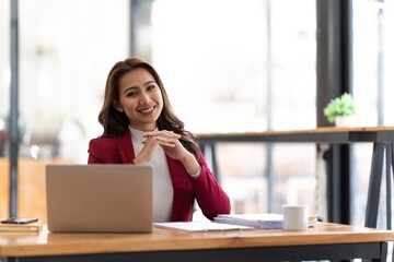 Obraz premium Young businesswoman working at her laptop and going over paperwork while sitting at a desk in an office