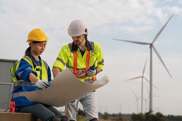 Male and female engineer in uniform with helmet safety holding blueprint and discussing to inspection and maintenance of wind turbine in wind farm to generate electrical energy, Renewable power energy