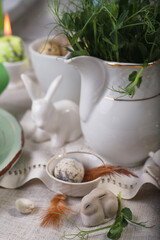 Easter table setting with fresh green plants, feathers, easter bunny and quail egg on white table.