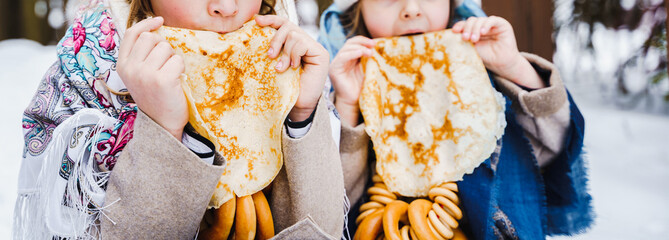 Russian national festival Maslenitsa, shrovetide. Little cute beautiful girls in headscarf eat big tasty pancakes, have fun on winter pancake holiday week. Traditional folf slavic street carnival