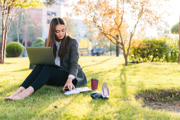 Woman freelancer in casual clothes sitting on the grass at the park writing her notes in her notebook with her computer on her legs. Freelance work with