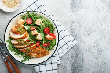 Fresh salad with grilled chicken fillet, arugula, tomatoes, cucumber and pesto sauce, sesame seeds with olive oil in white bowl on light slate background. Healthy lunch menu. Diet food. Top view.