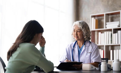 Woman senior doctor is Reading Medical History of Female Patient and Speaking with Her During Consultation in a Health Clinic. Physician in Lab Coat Sitting Behind a Laptop in Hospital Office