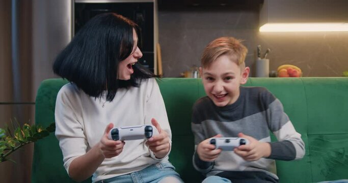 Young smilling woman and son playing video game together in moderm kitchen apartment enjoying indoor activity. Online entertainment and leisure activity concept.